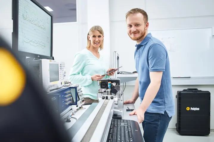 Two technicians smiling in a laboratory environment with Polytec vibration measurement equipment and monitors displaying technical data.