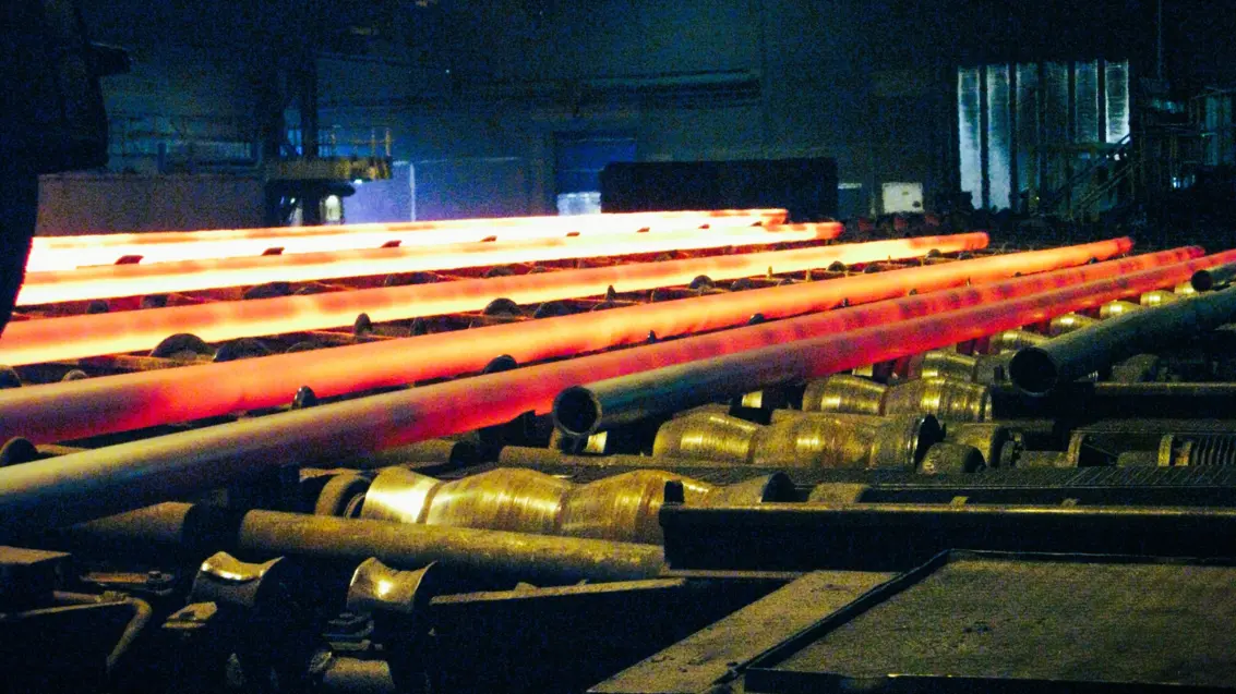 Glowing hot steel billets moving on industrial rollers in a dimly lit steel manufacturing facility.